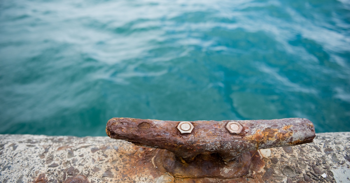 A close-up shot of an old, rusted dock cleat on the stone edge of a dock. The water is bright blue and clear.