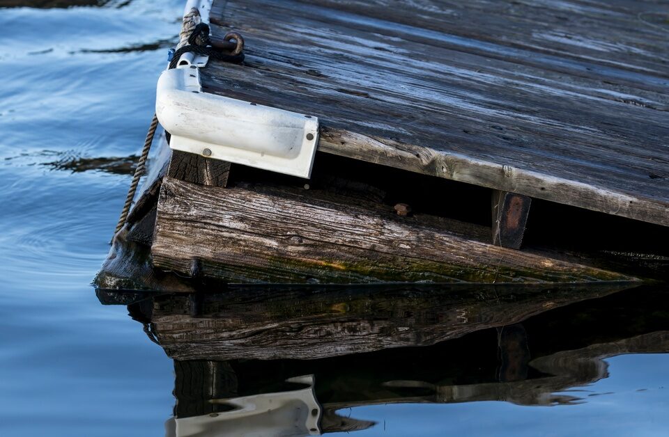 A corner of an old, wooden dock tilting in the water. The dock has protective covers on the corners.