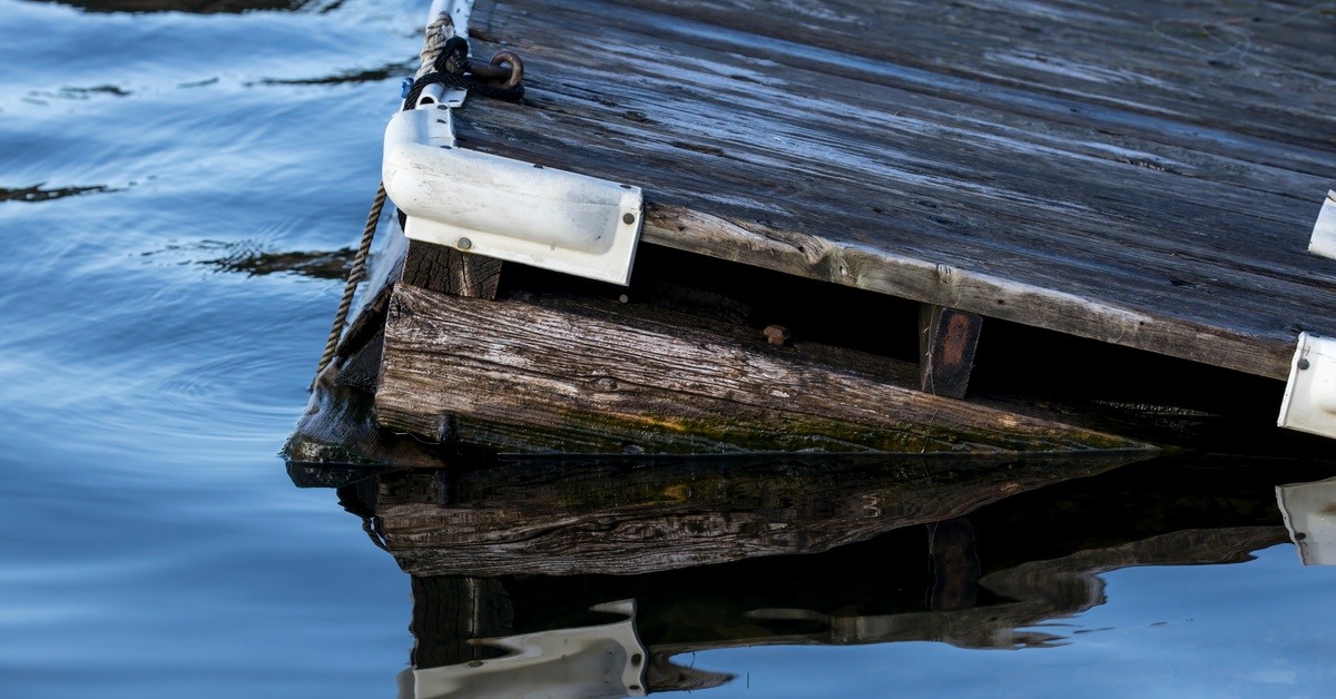 A corner of an old, wooden dock tilting in the water. The dock has protective covers on the corners.