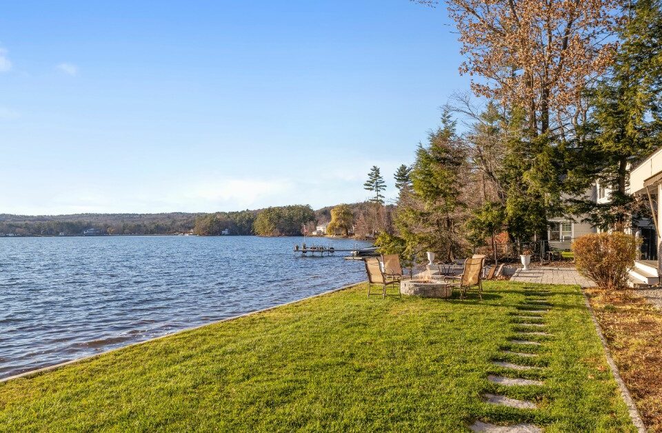 The back view of a modern, lakefront home with patio furniture, a fire pit, and a dock in the distance.