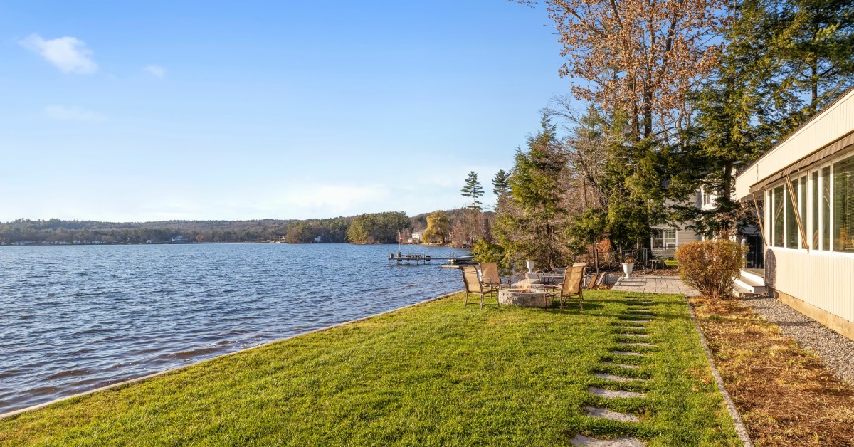 The back view of a modern, lakefront home with patio furniture, a fire pit, and a dock in the distance.