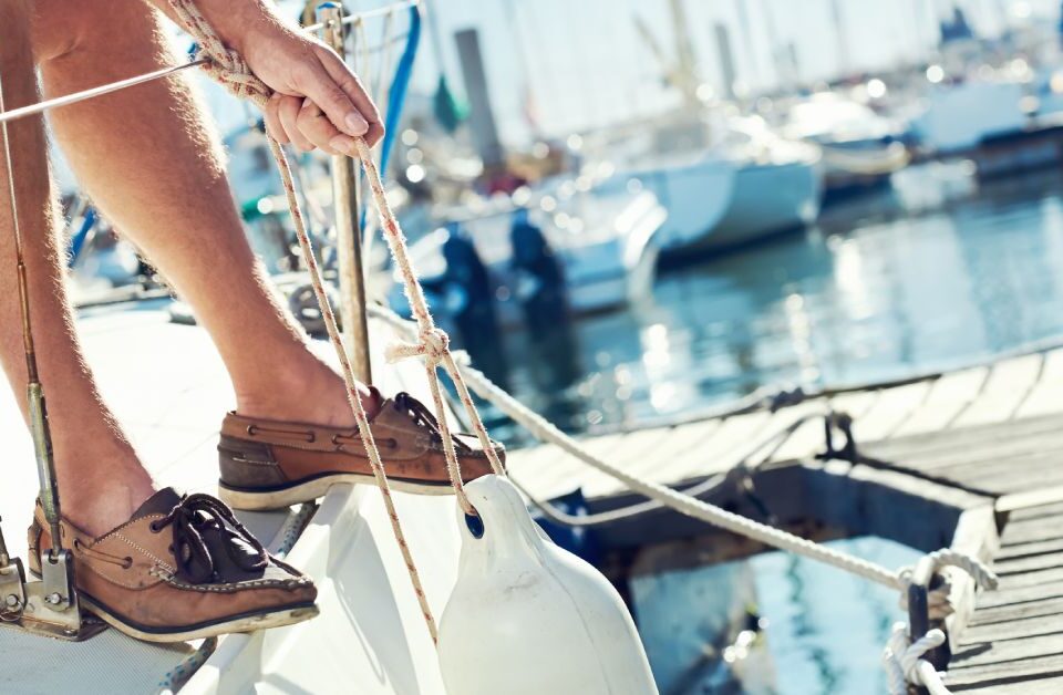 A person wearing boat shoes is standing on the edge of a boat and tying a line around a boat cleat.
