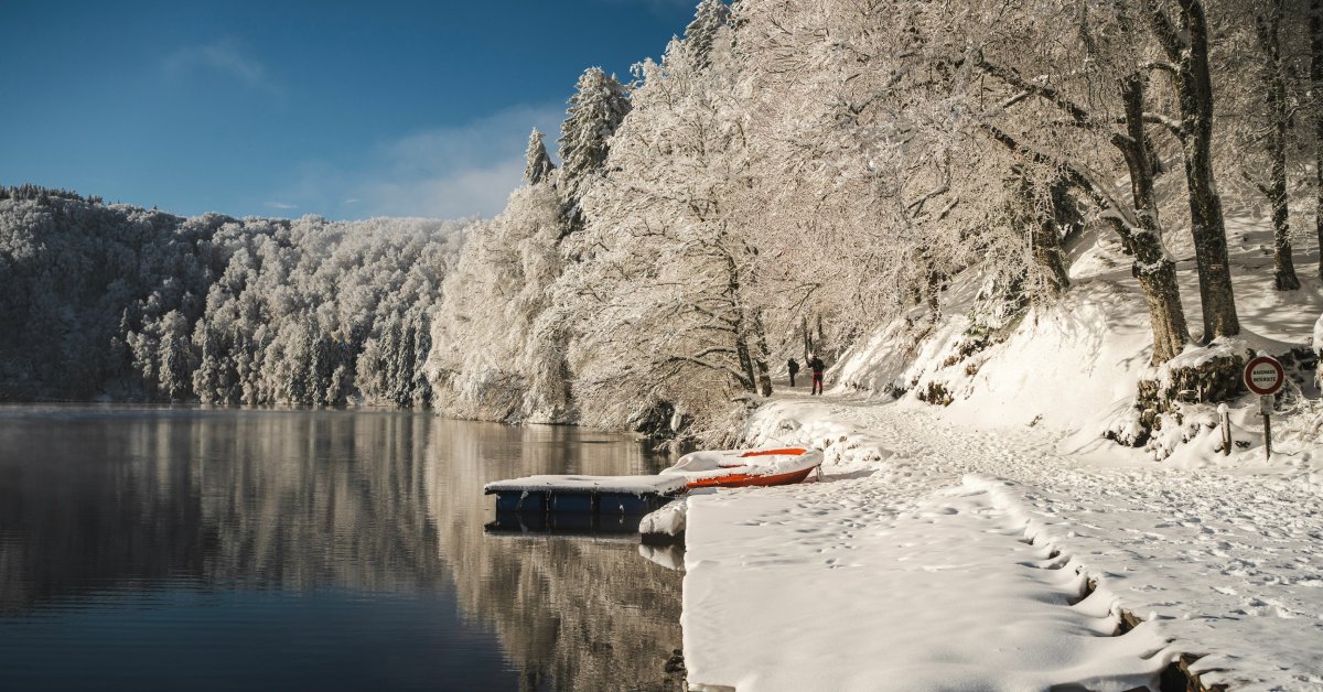 A still lake at the edge of the forest with a small dock and a red inflatable raft on the shore. Everything is blanketed in snow.