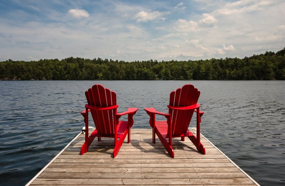 Two red Adirondack chairs sit next to each other on the edge of a dock, facing a lake on a partly cloudy day.