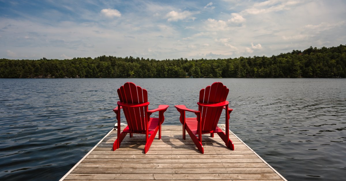Two red Adirondack chairs sit next to each other on the edge of a dock, facing a lake on a partly cloudy day.