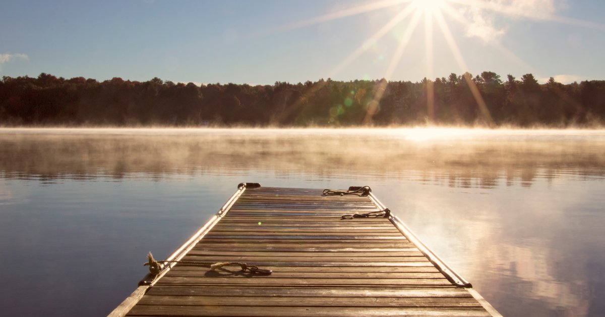 A wooden dock jutting into a lake with a forest in the distance. The sun is creating a glare in the upper right corner.