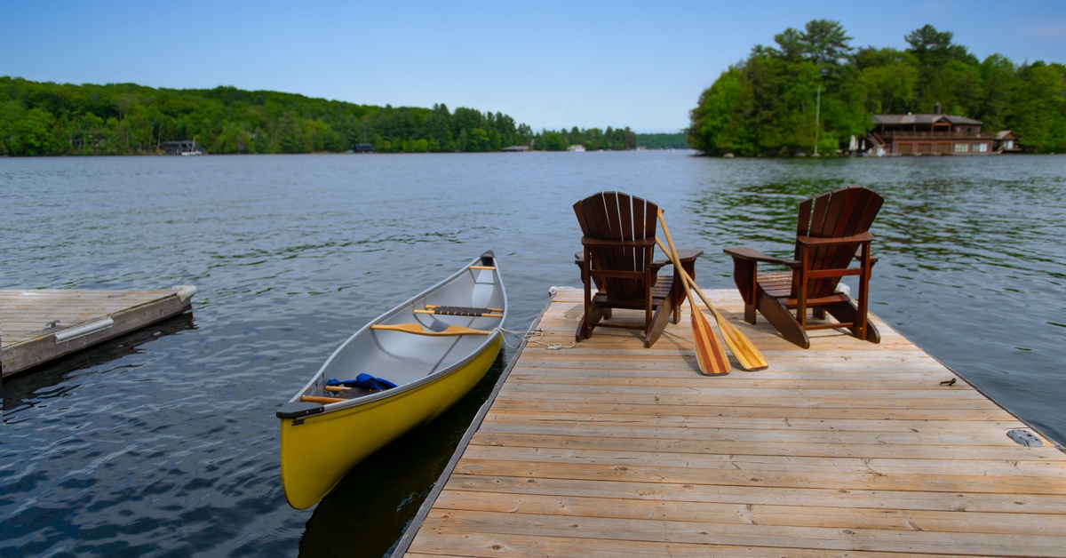 Two small wooden lakeside docks. The one in focus has two brown Adirondack chairs and a canoe tied to the dock.