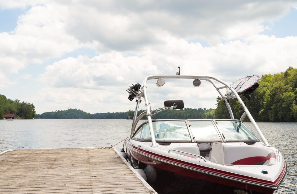 A red and white wakeboard boat tied by two anchor points to a small wooden dock. There are lakeside homes in the background.
