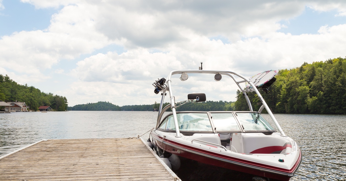 A red and white wakeboard boat tied by two anchor points to a small wooden dock. There are lakeside homes in the background.