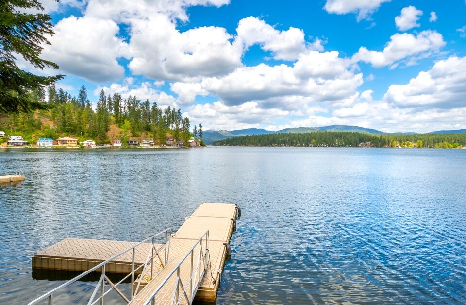 A large lake with a small dock under a cloudy blue sky, with homes on the shoreline and rolling mountains in the distance.