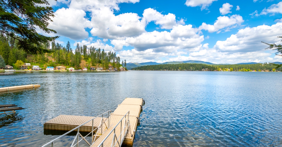 A large lake with a small dock under a cloudy blue sky, with homes on the shoreline and rolling mountains in the distance.