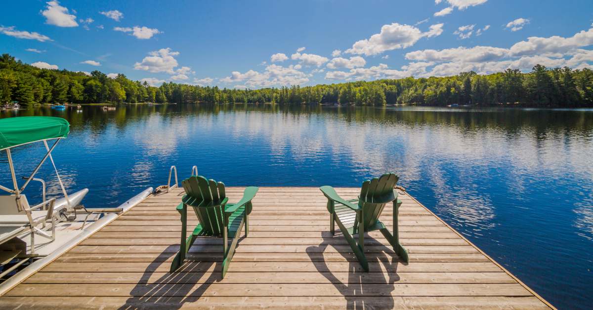 Two green Adirondack chairs at the end of a dock, looking over a beautiful lake; a small boat is attached.