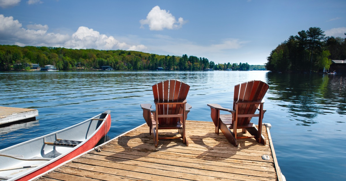 Two beach chairs set up at the end of a dock to look at the lake and forest beyond. Next to the dock is a small rowboat.