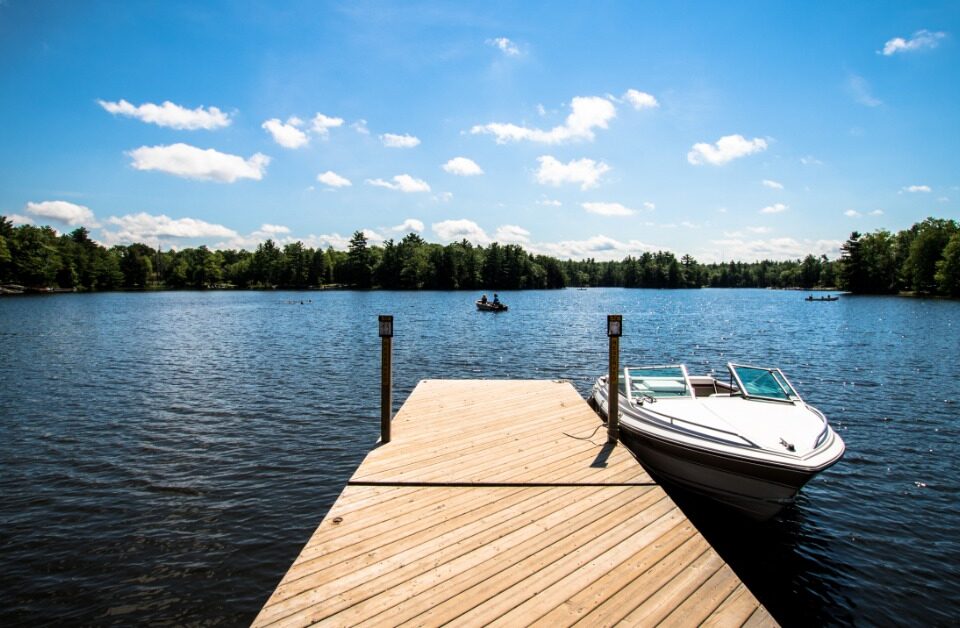 A wooden dock has a small boat tied to the end of it. The dock sits on a clear lake, surrounded by trees.
