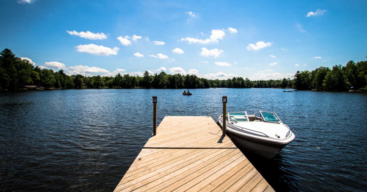 A wooden dock has a small boat tied to the end of it. The dock sits on a clear lake, surrounded by trees.