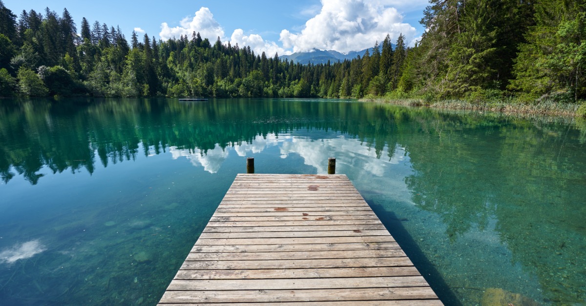 A short, wooden dock overlooking a clear lake. Beyond the lake lies a lush forest and a clear blue sky.