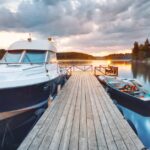 A wooden pier on a lake at sunset. There are two boats attached to the dock, one large and one small.