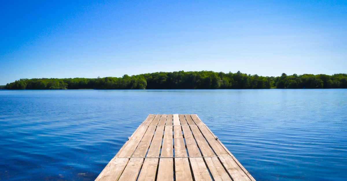 A wooden dock overlooking a clear lake. The sky above is clear, and there are trees on the other side of the lake.