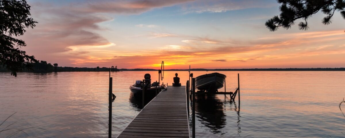 A young woman sitting at the end of a dock watching the sun set. There are two boats attached to the dock.