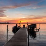 A young woman sitting at the end of a dock watching the sun set. There are two boats attached to the dock.