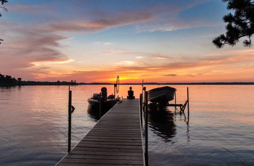 A young woman sitting at the end of a dock watching the sun set. There are two boats attached to the dock.