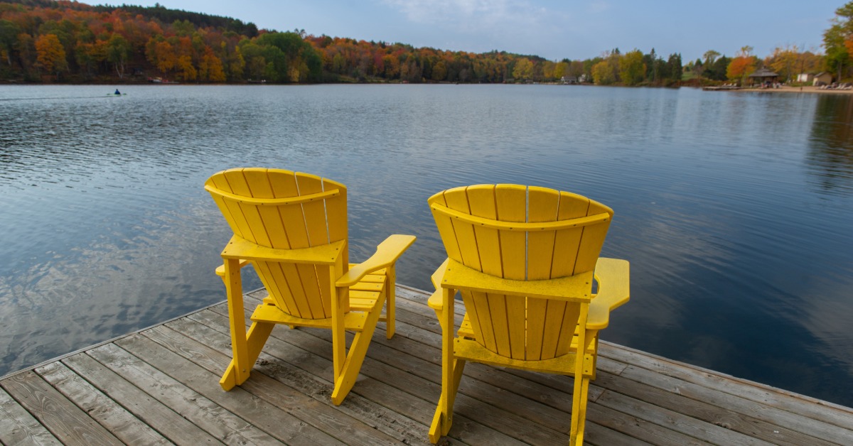 Two yellow chairs sitting on the corner of a dock, looking out over a clear, calm lake with trees on the other side.