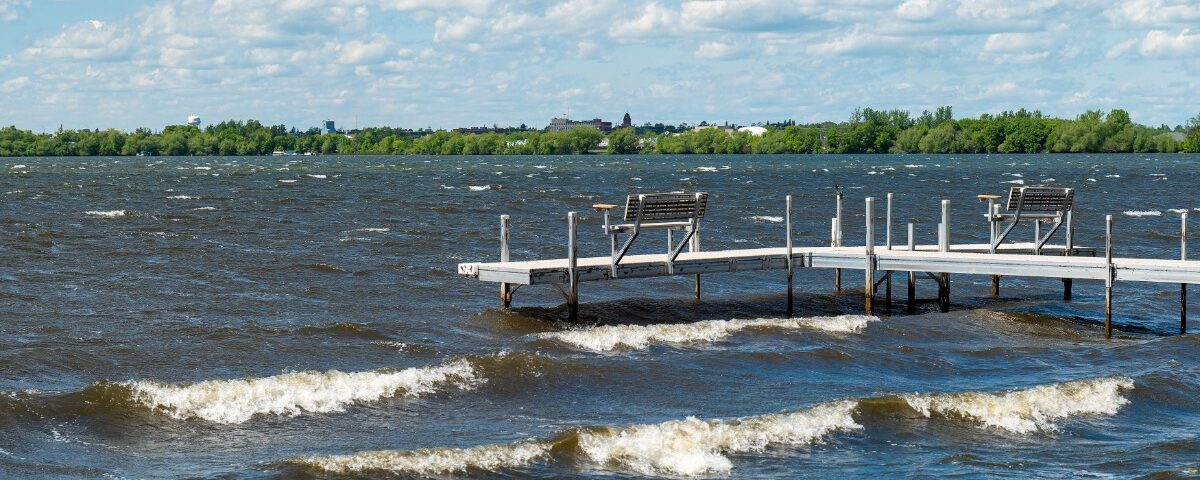 A long boat dock with benches looking out at the water. The water has a variety of waves rolling towards the dock.