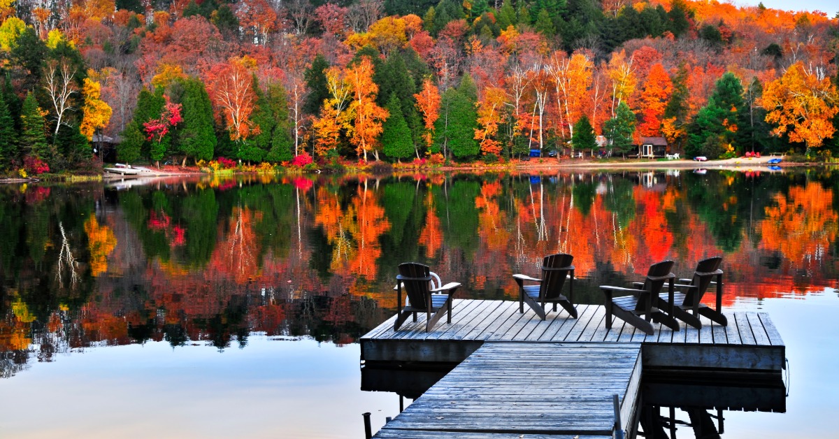 A wooden dock with chairs set up to look out on a lake. Across the lake, multicolored trees are reflected in the water.