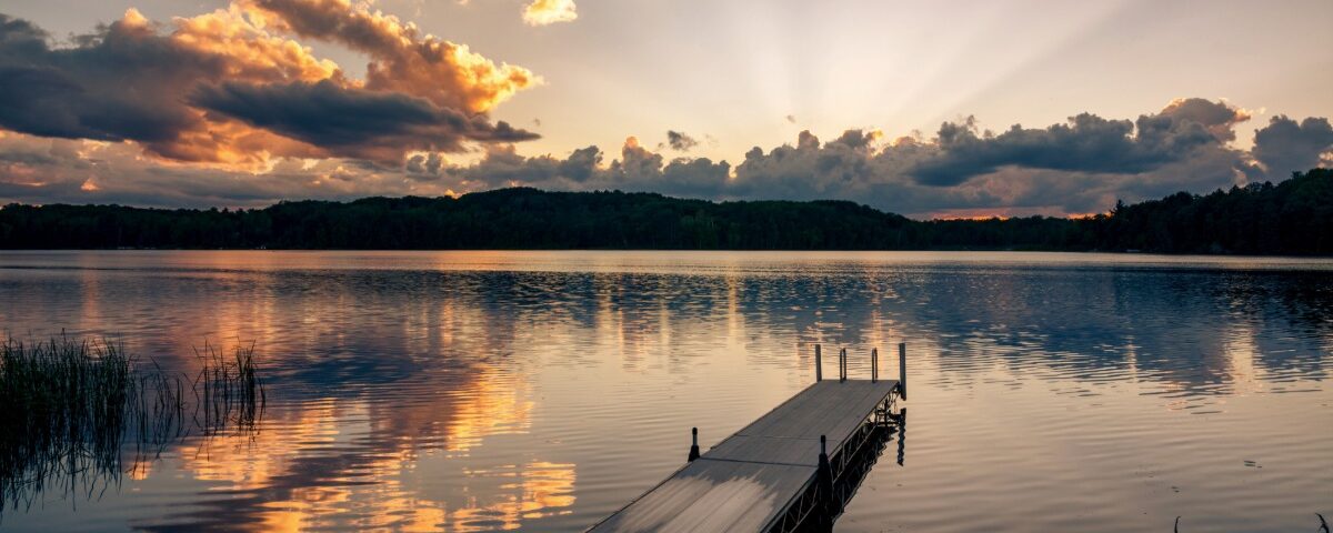 A small dock jutting out onto a clear lake with nothing on it. The water is reflecting the sunset overhead.