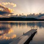A small dock jutting out onto a clear lake with nothing on it. The water is reflecting the sunset overhead.