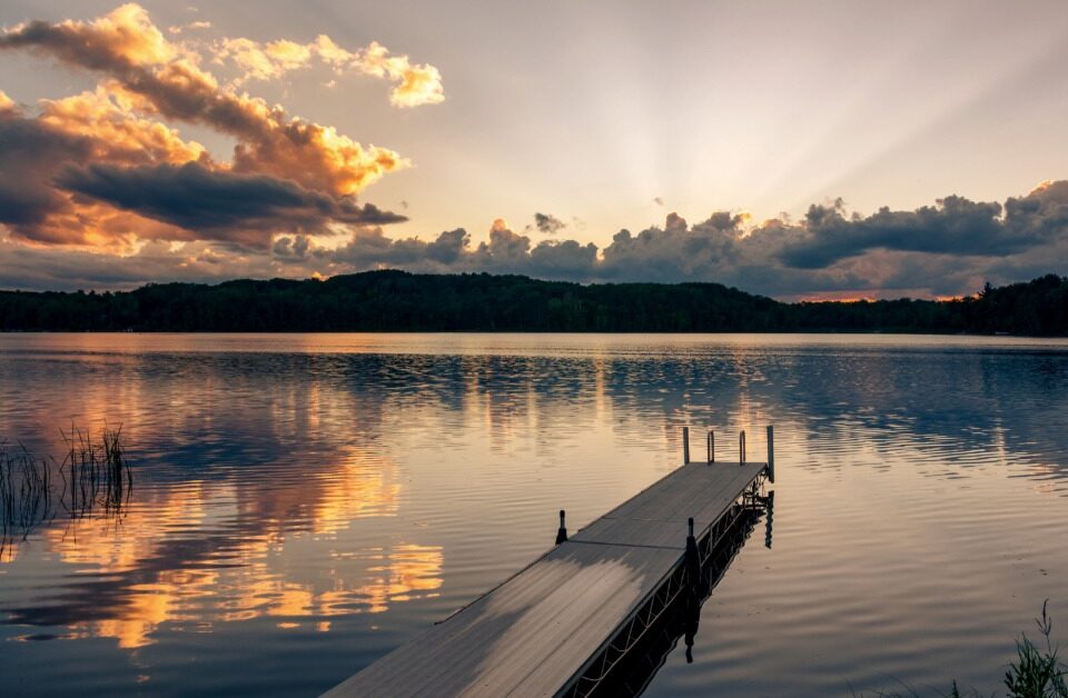 A small dock jutting out onto a clear lake with nothing on it. The water is reflecting the sunset overhead.