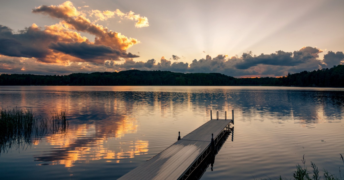A small dock jutting out onto a clear lake with nothing on it. The water is reflecting the sunset overhead.
