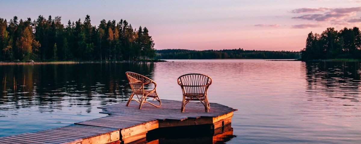 Two wooden chairs on a wooden deck looking out over a lake. The deck leads to a small circle with the chairs.