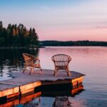 Two wooden chairs on a wooden deck looking out over a lake. The deck leads to a small circle with the chairs.