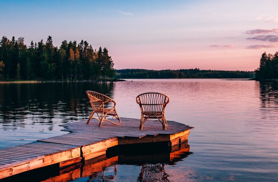 Two wooden chairs on a wooden deck looking out over a lake. The deck leads to a small circle with the chairs.