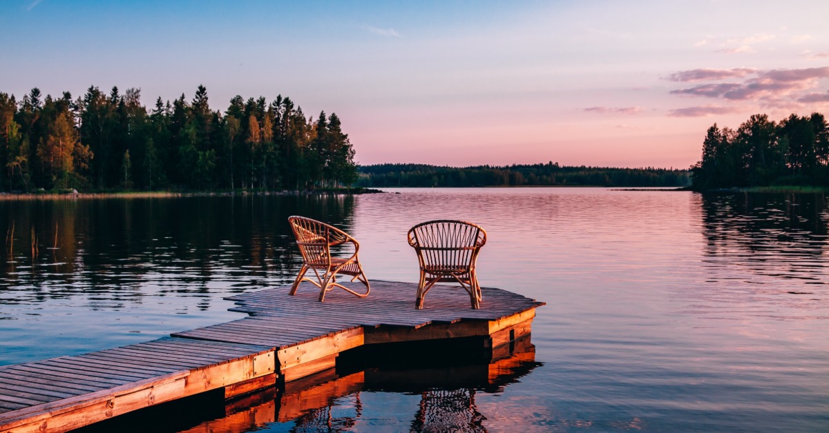 Two wooden chairs on a wooden deck looking out over a lake. The deck leads to a small circle with the chairs.