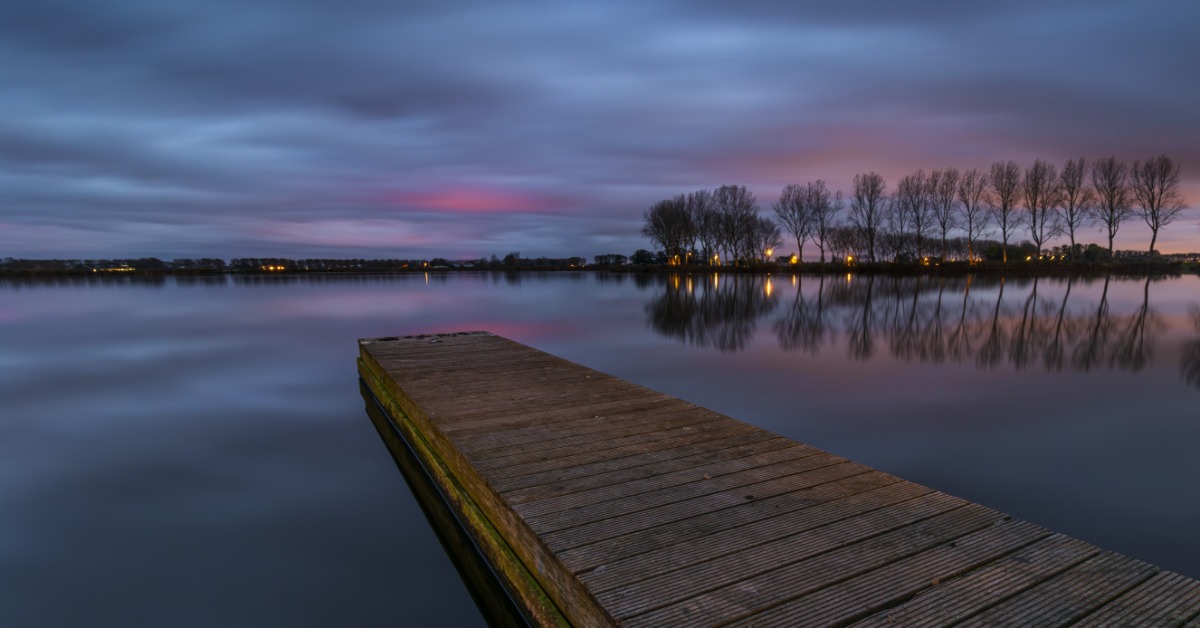 A long, wooden deck sitting on a calm lake at sunset. There are some trees in the distance that reflect on the water.