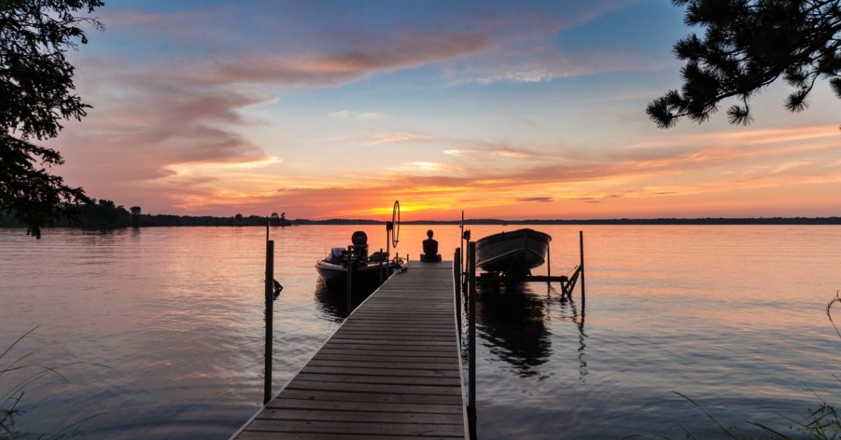 A person sitting on the edge of a dock, looking at the sunset over the lake. There are two boats connected to the dock.