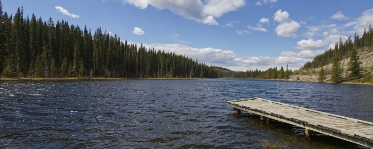A wooden dock extending into a rippled lake surrounded by evergreen trees, rocky shore, blue sky, and white clouds.