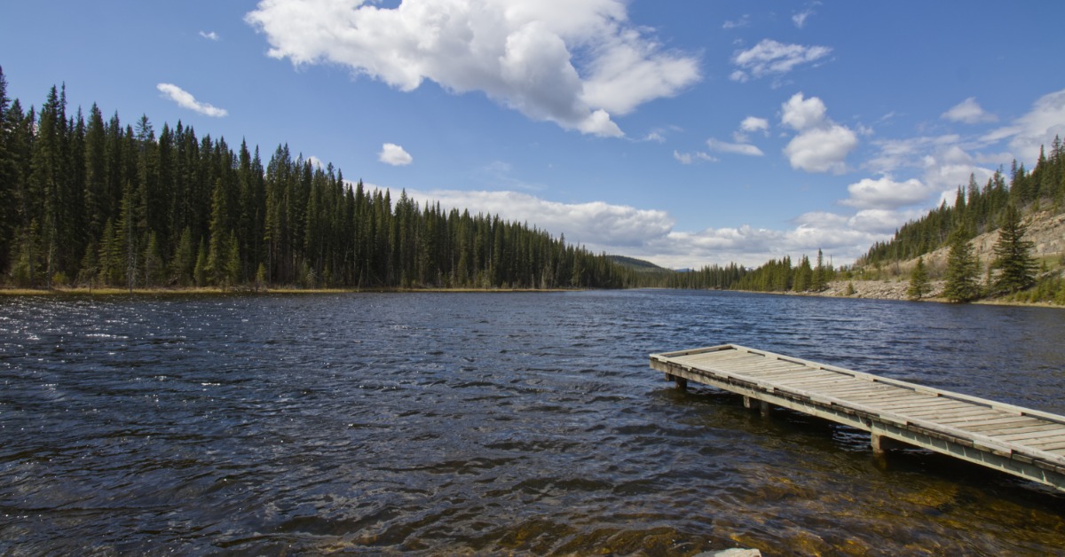 A wooden dock extending into a rippled lake surrounded by evergreen trees, rocky shore, blue sky, and white clouds.