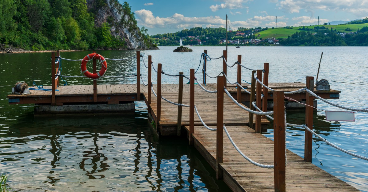 A wooden pier with rope railings and a lifebuoy extending into a lake with forested hills in the distance.