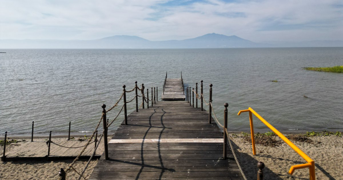 A rustic wooden pier extending into calm lake water, with sandy shore, distant hazy mountains, and cloudy sky.