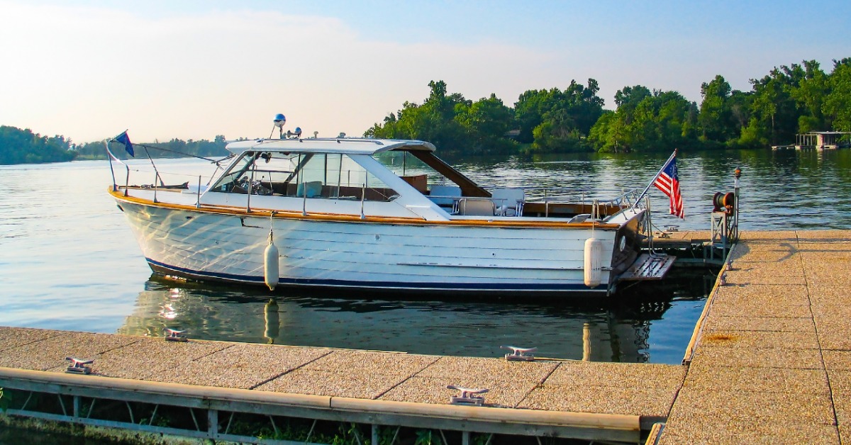 A wooden boat with an American flag moored beside a dock on a calm lake, with trees reflected in the water.
