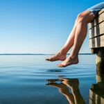 A person sitting on a wooden dock with feet over calm lake water, with trees and a pale sky in the distance.