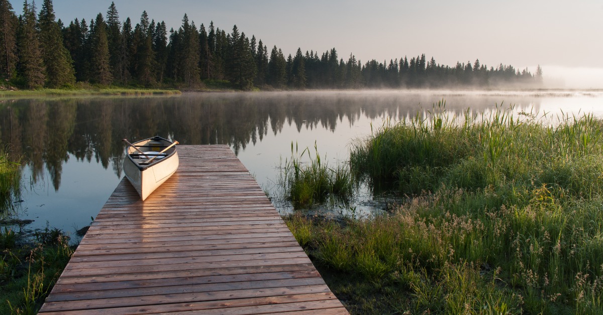 A canoe resting on a wooden dock beside a misty lake, with trees, grass, and sunrise light over calm water.