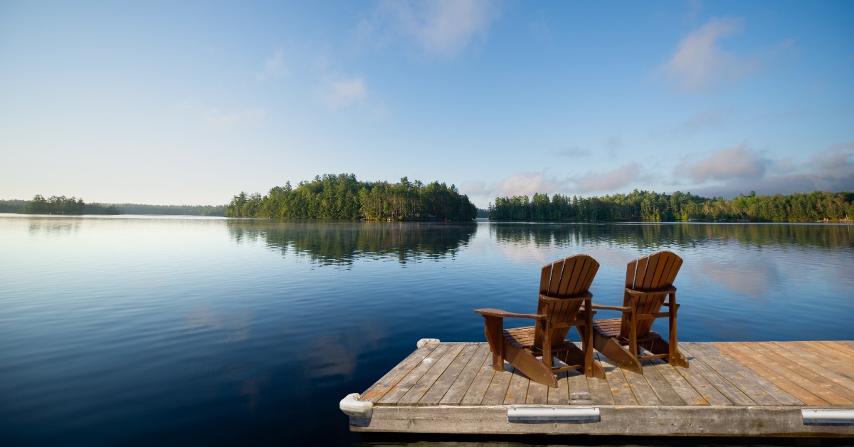 A wooden dock overlooking a calm lake with trees in the distance. There are two Adirondack chairs on the dock.