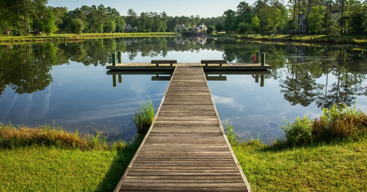 A wooden pier leading out onto a small lake or pond with two sides. The grass around the dock is bright green.